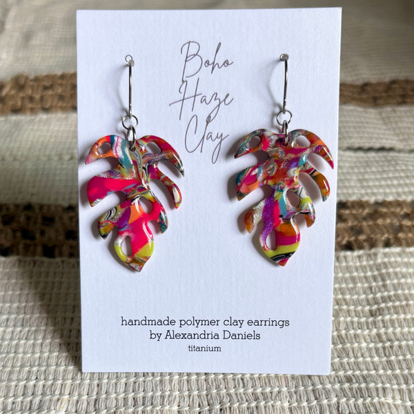 A pair of handmade polymer clay earrings by Alexandria Daniels, displayed on a white card. The earrings are shaped like tropical palm leaves and feature a vibrant, multicolored marbled pattern of pink, orange, yellow, green, and purple.
