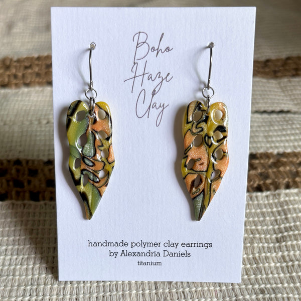 A pair of handmade polymer clay dangle earrings by Alexandria Daniels, displayed on a white card. The earrings feature a marbled pattern of olive green, terracotta orange, and black, suspended from silver fishhook wires.