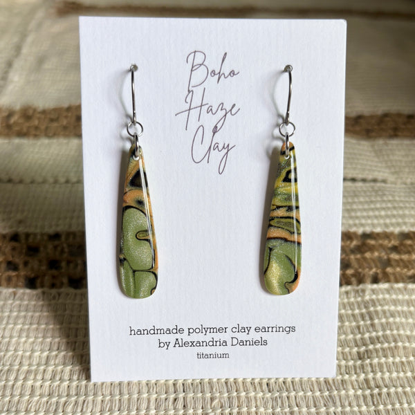 A pair of handmade polymer clay dangle earrings with a green, gold, and black marbled pattern, displayed on a white card labeled 'Boho Haze Clay' by Alexandria Daniels.