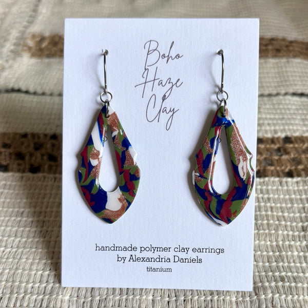 A pair of handmade polymer clay dangle earrings with a marbled pattern of blue, green, red, and white, displayed on a white card labeled 'Boho Haze Clay' by Alexandria Daniels.