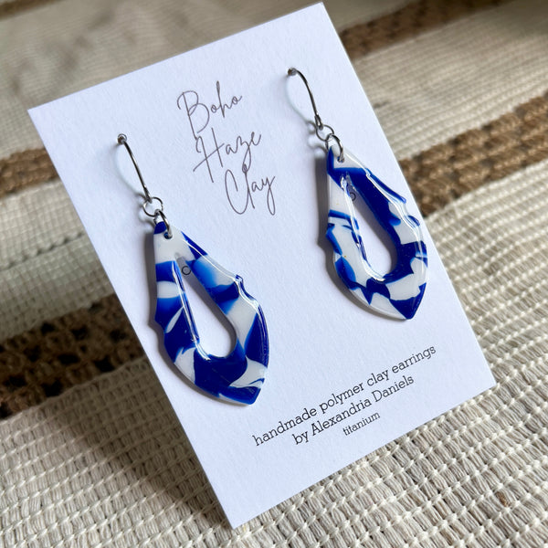 Blue and white marbled polymer clay dangle earrings displayed on a white card labeled 'Boho Haze Clay' by Alexandria Daniels, placed on a woven beige surface.