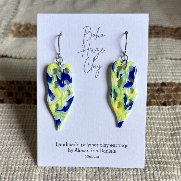 A pair of handmade polymer clay dangle earrings by Alexandria Daniels, featuring a blue, yellow, and lime green marbled pattern on a white display card labeled 'Boho Haze Clay'.