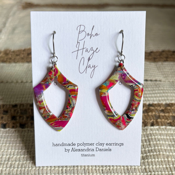 A pair of handmade polymer clay earrings by Alexandria Daniels, featuring a vibrant multicolor swirl pattern in pink, yellow, orange, and black, displayed on a white card labeled 'Boho Haze Clay'.