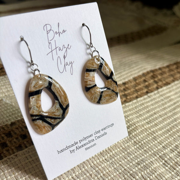 A pair of handmade polymer clay earrings displayed on a white card. The earrings are teardrop-shaped with a natural beige and brown marbled pattern and black veins, suspended from silver fishhook wires.