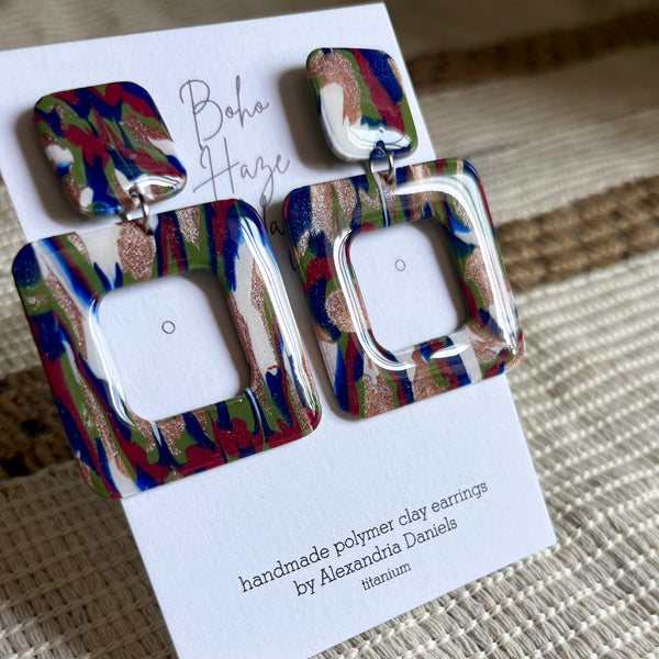 A pair of colorful square drop earrings displayed on a white branded card. The earrings feature a marbled pattern of blue, green, red, and gold glitter, with a hollow square cutout in the center.