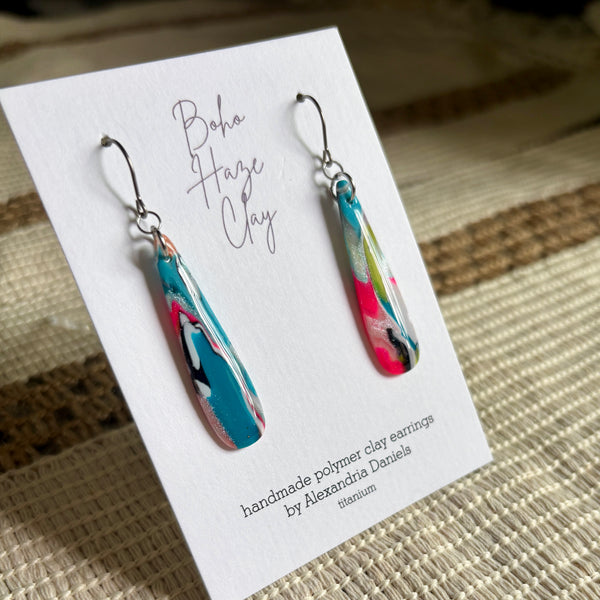 A pair of handmade teardrop-shaped polymer clay earrings displayed on a white branded card. The earrings feature a marbled swirl pattern of turquoise blue, pink, lime green, and black, suspended from silver fishhook wires.