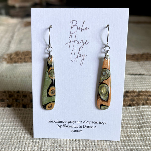 A pair of handmade polymer clay dangle earrings by Alexandria Daniels, displayed on a white card. The earrings feature organic, swirling patterns in olive green, terracotta orange, and black on a silver-tone hook setting.
