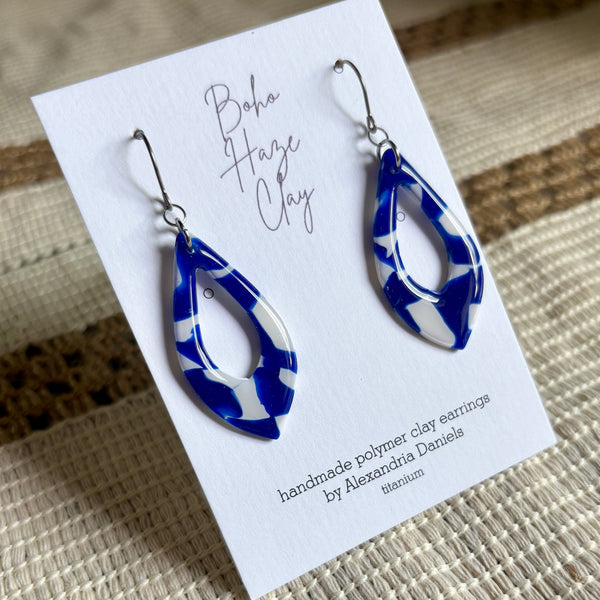 A pair of blue and white patterned polymer clay dangle earrings on a white display card, photographed on a woven beige surface. The card reads 'Boho Haze Clay' and 'handmade polymer clay earrings by Alexandria Daniels titlerium'.