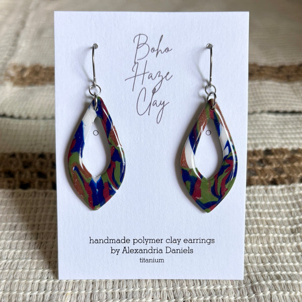 A pair of handmade polymer clay dangle earrings by Alexandria Daniels, displayed on a white card. The earrings feature a marbled pattern of blue, green, red, and white clay with silver fishhook wires.