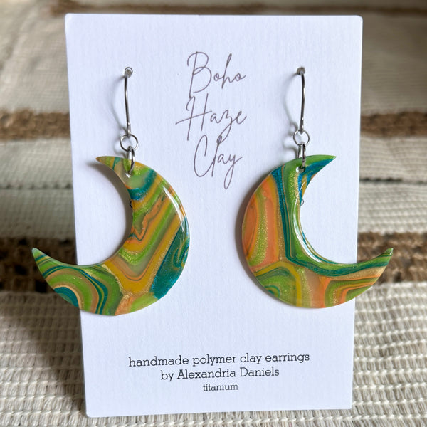 A pair of handmade crescent moon polymer clay earrings with a marbled green, yellow, and orange pattern, displayed on a white card that reads 'Boho Haze Clay' and 'handmade polymer clay earrings by Alexandria Daniels'.
