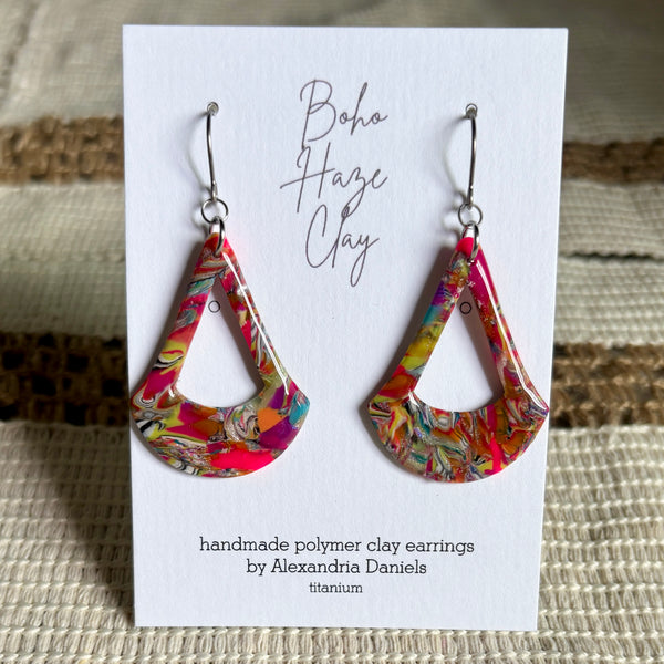 A pair of handmade polymer clay dangle earrings by Alexandria Daniels, displayed on a white card. The earrings feature a vibrant, multicolored swirl pattern in shades of pink, yellow, green, and blue, suspended from silver fishhook wires.
