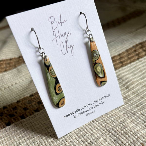 A pair of handmade polymer clay dangle earrings displayed on a white card. The earrings feature organic, wavy patterns in olive green, terracotta orange, and black, suspended from silver fishhook wires.