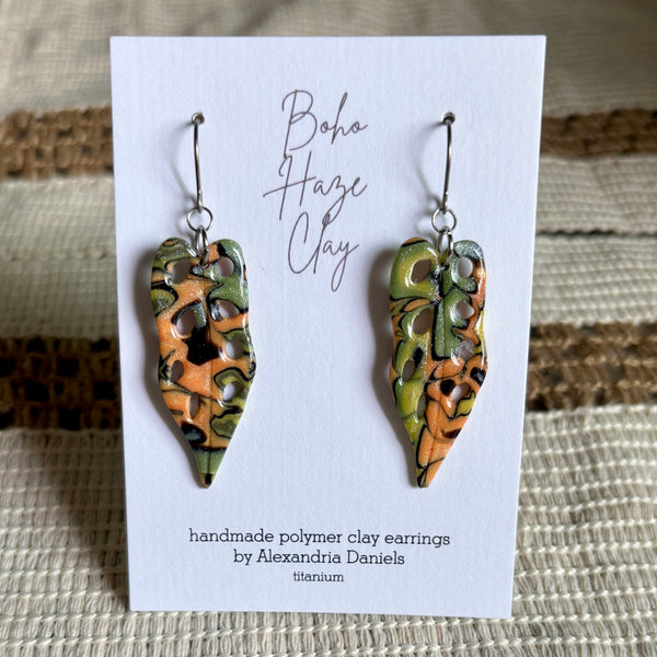 A pair of handmade polymer clay dangle earrings by Alexandria Daniels, displayed on a white card. The earrings feature an abstract organic pattern in orange, olive green, and black on a titanium wire base.