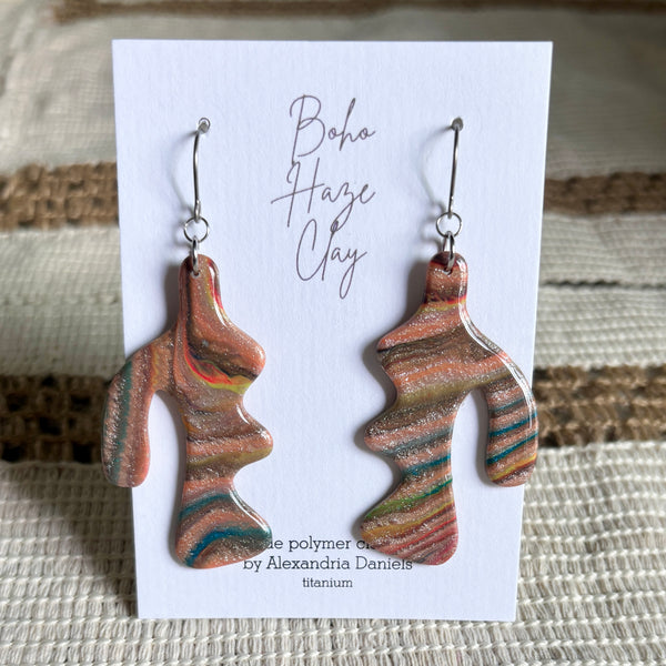 A pair of abstract sculptural dangle earrings displayed on a white backing card. The earrings have a marbled, fluid pattern of terracotta orange, brown, teal, and gold glitter, suspended from silver fishhook wires.