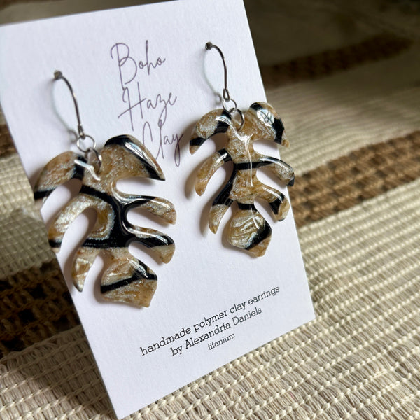 A pair of handmade polymer clay earrings shaped like monstera leaves, displayed on a white card that reads 'Boho Haze Clay' and 'handmade polymer clay earrings by Alexandra Daniels'. The earrings feature a marbled pattern of beige, black, and brown, with silver-toned fishhook wires.