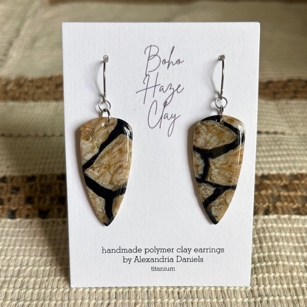 A pair of handmade polymer clay dangle earrings with a polished gold and black patterned design, displayed on a white card that reads 'Boho Haze Clay' by Alexandria Daniels.