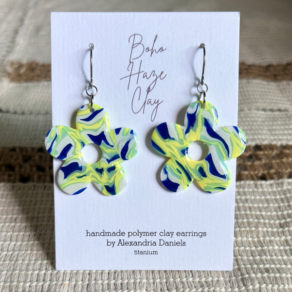 A pair of handmade polymer clay earrings shaped like flowers, featuring a marbled swirl pattern of bright yellow, green, and navy blue. The earrings are displayed on a white card labeled 'Boho Haze Clay' by handmaker Alexandria Daniels, set against a textured beige woven background.