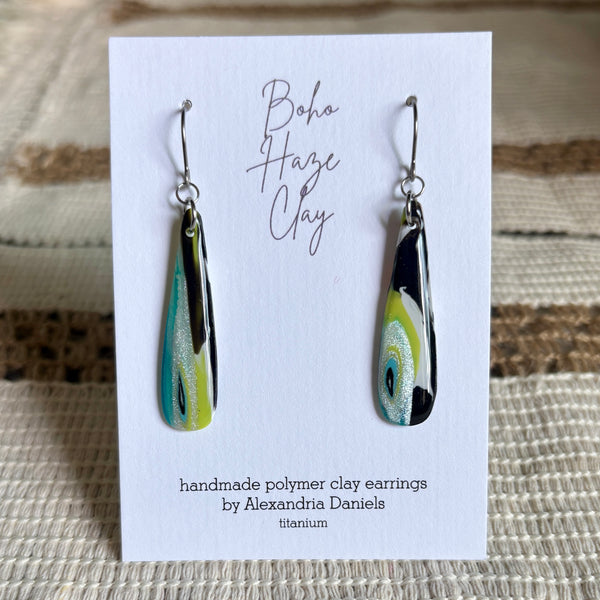 A pair of handmade polymer clay dangle earrings by Alexandria Daniels, displayed on a white card. The earrings feature a marbled pattern of black, lime green, and white with silver glitter, suspended from silver hooks.