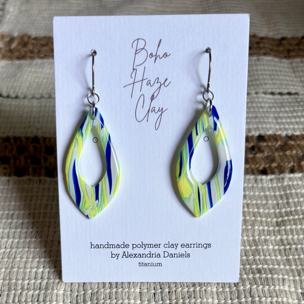 Teardrop-shaped polymer clay earrings with a blue, yellow, and white marbled pattern, displayed on a white card labeled 'Boho Haze Clay' by Alexandria Daniels.