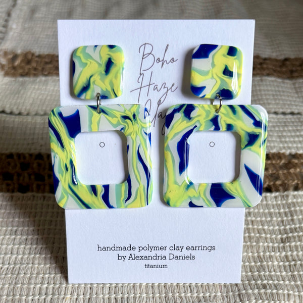 A pair of handmade polymer clay earrings displayed on a white card. The earrings feature a marbled swirl pattern of bright yellow, navy blue, and white.