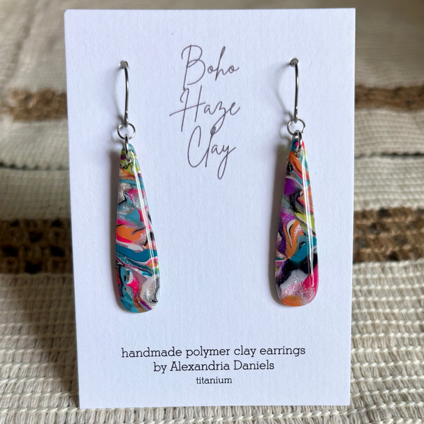 A pair of colorful teardrop-shaped polymer clay dangle earrings displayed on a white card. The earrings feature a vibrant marbled swirl pattern with pink, orange, teal, yellow, and black, suspended from silver fishhook wires.