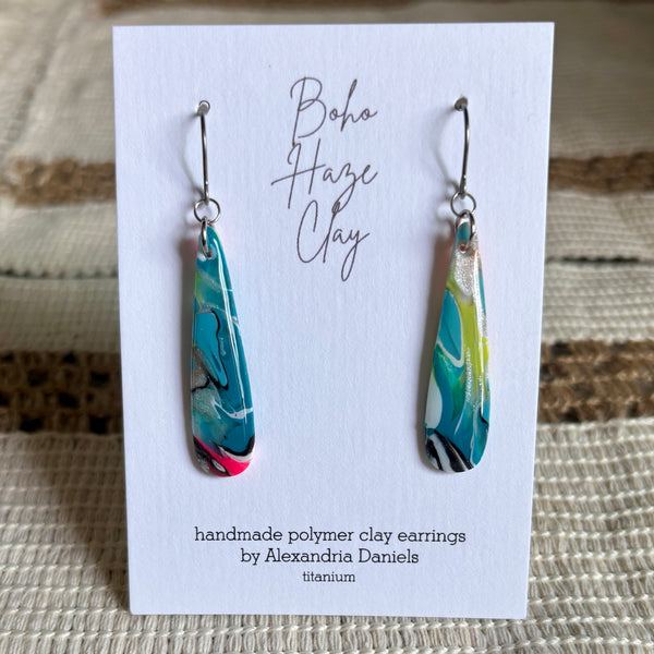 A pair of handmade polymer clay dangle earrings by Alexandria Daniels, displayed on a white backing card. The earrings are made of titanium with a marbled pattern of teal blue, lime green, yellow, pink, and black swirls.