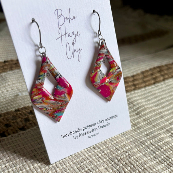 A pair of colorful geometric polymer clay dangle earrings displayed on a white branded card. The earrings feature a marbled pattern of hot pink, orange, yellow, and teal on a titanium base, with silver fishhook wires.