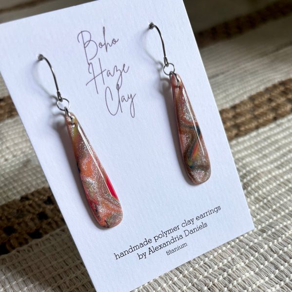 A pair of teardrop-shaped polymer clay earrings with a swirling orange, red, and green pattern and metallic gold flecks, displayed on a white card labeled 'Boho Haze Clay' by Alexandra Daniels.