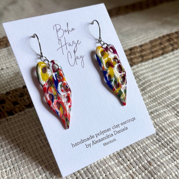 A pair of colorful handmade polymer clay earrings displayed on a white card. The earrings have a marbled, abstract pattern of yellow, red, blue, green, and white, suspended from silver fishhook wires.