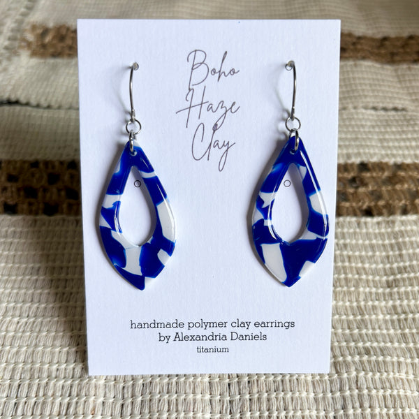 A pair of handmade polymer clay dangle earrings with a blue and white marbled pattern, displayed on a white card that reads 'Boho Haze Clay' and 'handmade polymer clay earrings by Alexandria Daniels'. The card is placed on a textured beige woven mat.