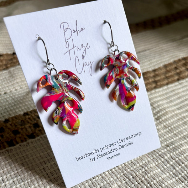 A pair of colorful polymer clay dangle earrings displayed on a white branded card. The earrings have a vibrant, multicolored marbled pattern with pink, yellow, green, and blue tones, suspended from silver fishhook wires.