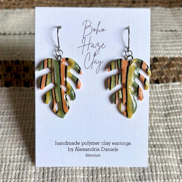 A pair of handmade polymer clay dangle earrings shaped like monstera leaves, displayed on a white card. The earrings feature a metallic titanium base with hand-painted green, orange, and black patterns.