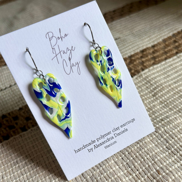A pair of handmade polymer clay earrings displayed on a white card. The earrings are shaped like stylized leaves with a marbled pattern of bright yellow, deep blue, and light green, suspended from silver fishhook wires.