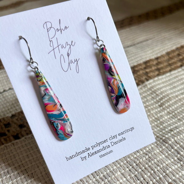 A pair of handmade polymer clay dangle earrings displayed on a white card. The earrings feature a vibrant marbled pattern of turquoise, pink, orange, black, and white, suspended from silver fishhook wires.