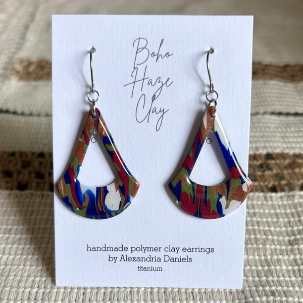 A pair of handmade polymer clay dangle earrings by Alexandria Daniels, displayed on a white card. The earrings feature a marbled pattern of red, blue, green, and white clay with a small silver bead at the center and silver fishhook ear wires.