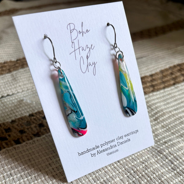 A pair of teardrop-shaped polymer clay earrings with a marbled pattern of teal, green, white, and pink, displayed on a white card that reads 'Boho Haze Clay' by Alexandra Daniels. The card is placed on a woven beige surface.