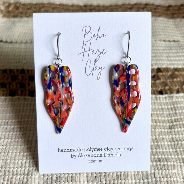 A pair of colorful handmade polymer clay dangle earrings displayed on a white card. The earrings feature an abstract marbled pattern in red, orange, yellow, blue, and white, suspended from silver fishhook wires.