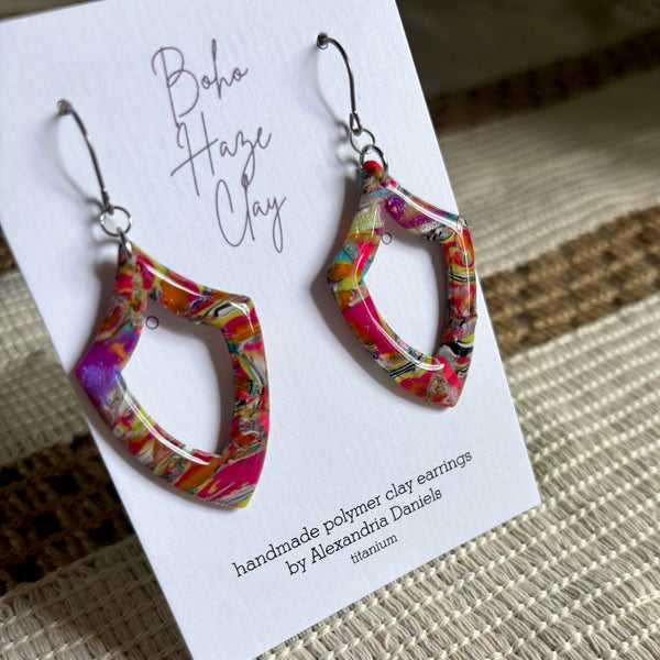 A pair of colorful open-loop polymer clay dangle earrings displayed on a white card that reads 'Boho Haze Clay' and 'handmade polymer clay earrings by Alexandria Daniels.' The earrings have a glossy marbled pattern with bright pink, orange, yellow, green, and purple tones, hanging from silver fishhook wires.
