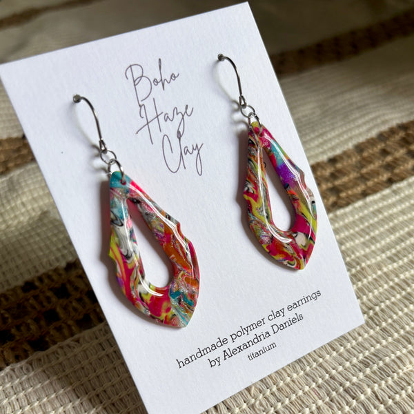 A pair of colorful teardrop-shaped polymer clay earrings displayed on a white card. The earrings feature a vibrant marbled pattern with pink, red, yellow, green, and blue swirls, suspended from silver fishhook wires.