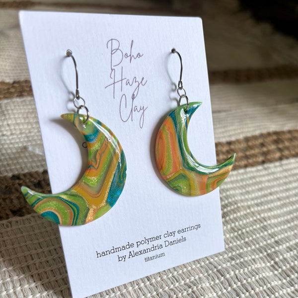 A pair of handmade crescent moon polymer clay earrings with a marbled pattern of green, yellow, and orange, hanging from silver fishhook wires on a white display card. The card is placed on a woven beige fabric background.