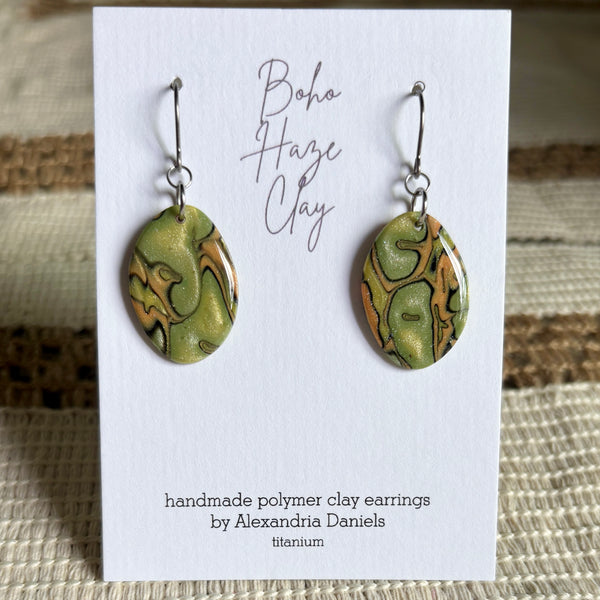 A pair of handmade polymer clay dangle earrings by Alexandria Daniels, displayed on a white card. The earrings feature organic marbled swirls of olive green, terracotta orange, and metallic gold with silver fishhook wires.