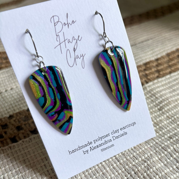 A pair of bohemian polymer clay earrings displayed on a white branded card. The earrings feature a glossy, marbled pattern of black, purple, green, and blue with a metallic sheen, suspended from silver fishhook wires.