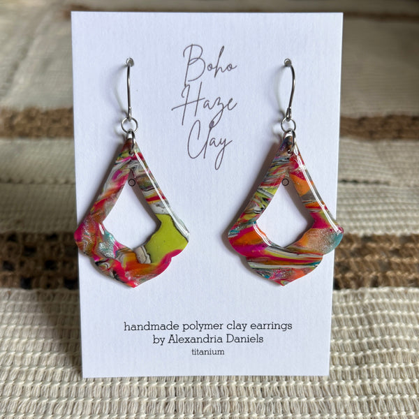 A pair of colorful handmade polymer clay dangle earrings displayed on a white card. The earrings have a marbled, geometric design with bright pink, orange, yellow, green, and white colors.