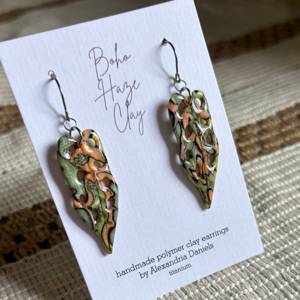 A pair of handmade polymer clay earrings displayed on a white card. The earrings have a textured, organic pattern in olive green, terracotta orange, and cream, suspended from silver fishhook wires.