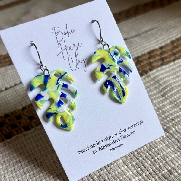 A pair of handmade polymer clay earrings shaped like monstera leaves, featuring a marbled swirl of yellow, blue, and white. They are displayed on a white card that reads 'Boho Haze Clay' and 'handmade polymer clay earrings by Alexandria Daniels', placed on a woven beige surface.