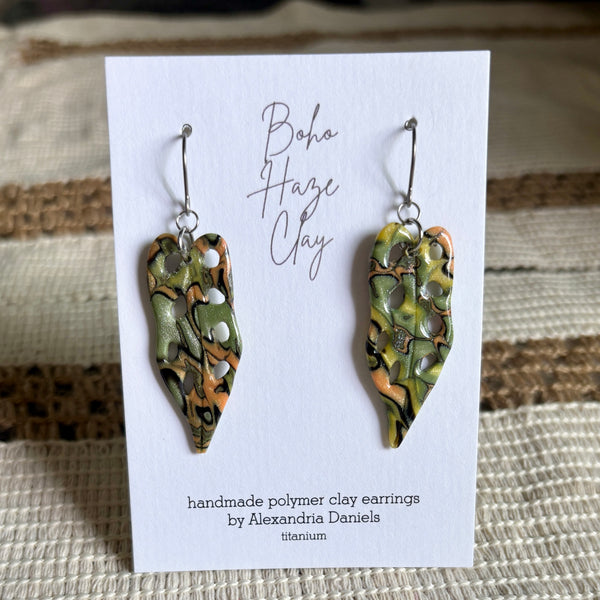 A pair of handmade polymer clay dangle earrings displayed on a white card. The earrings feature a marbled pattern of olive green, terracotta orange, and black, with small silver metallic dots.