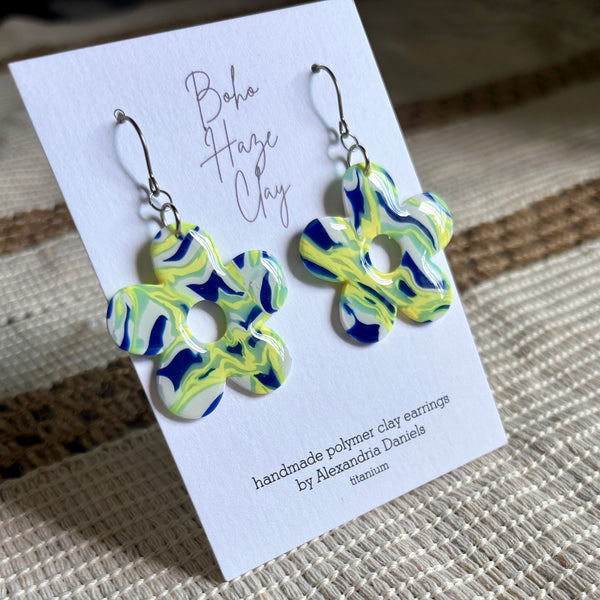 A pair of flower-shaped polymer clay earrings in a marbled blue, yellow, and white pattern, displayed on a white card that reads 'Boho Haze Clay' and 'handmade polymer clay earrings by Alexandria Daniels'.
